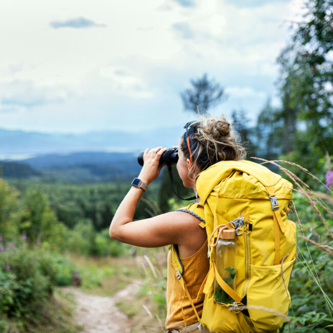 Rear view of woman hiker with backpack on a hiking trip in nature, using binoculars.