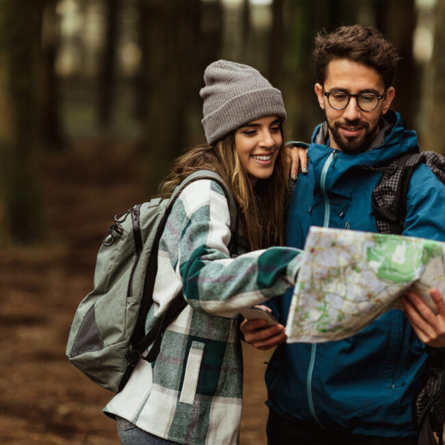 Cheerful young caucasian couple in jackets enjoy travel, vacation, look at map for route in forest